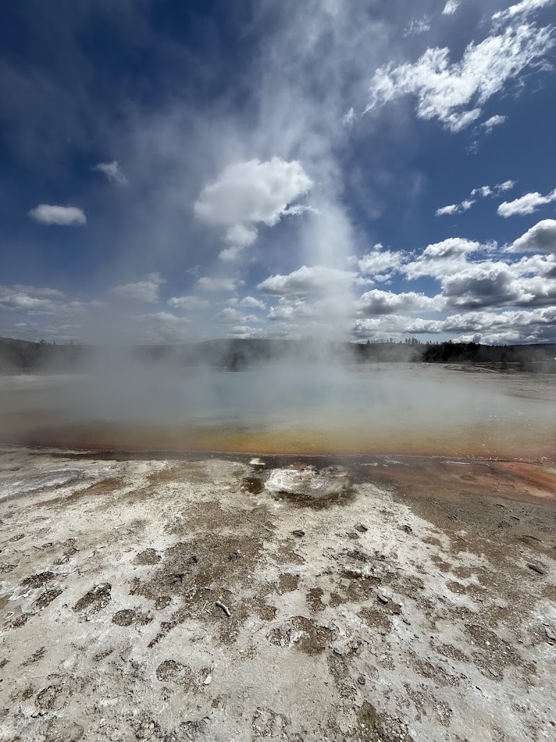 Steam rises from a rainbow-colored geothermal pool with orange-brown crust, under a blue sky over Yellowstone National Park.