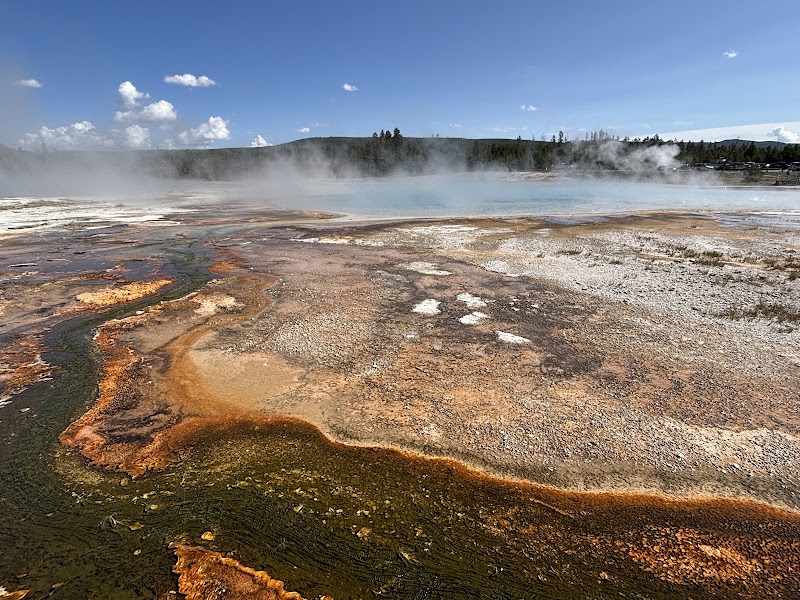 Steam rises above a blue Sunset Lake geothermal pool in Yellowstone National Park, with vivid orange, brown, and green mineral terraces in the sunlit foreground.