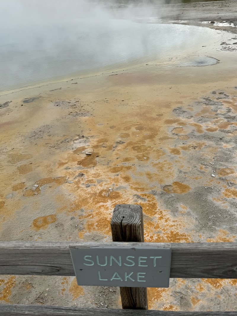 Wooden railing frames a steaming thermal pool with orange bacterial mats along the shore, Sunset Lake, Yellowstone National Park.