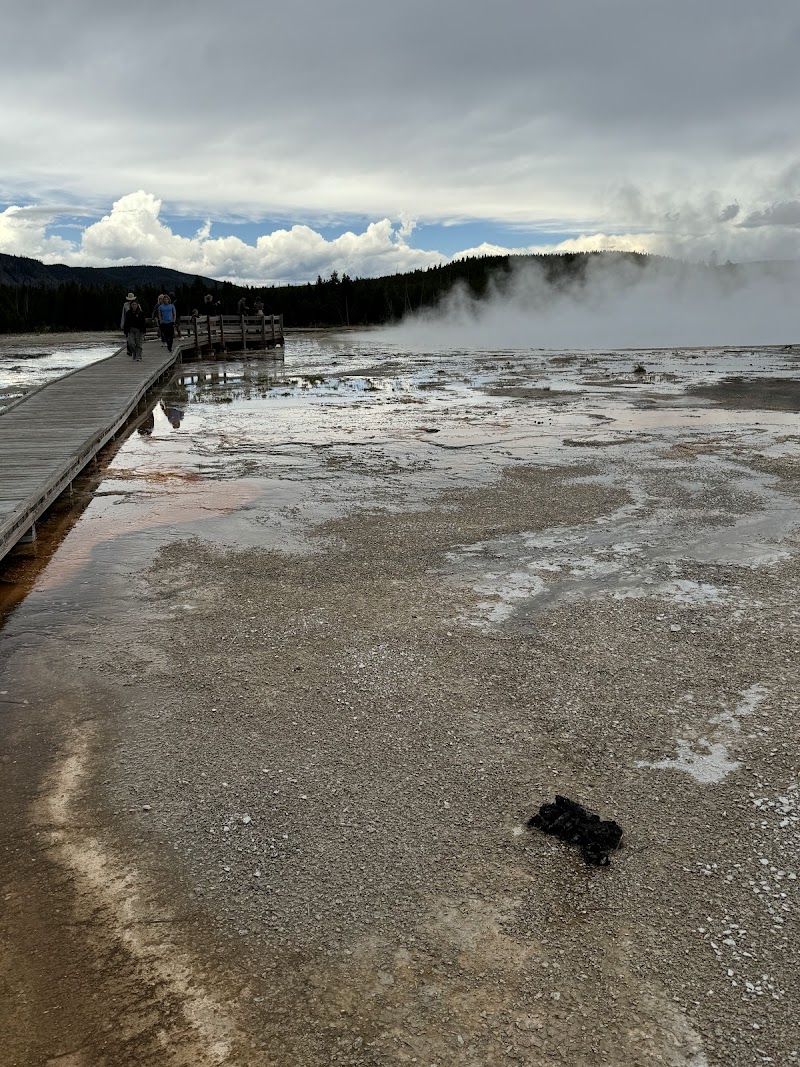 Wooden boardwalk winds through a steaming geothermal field at Sunset Lake, Yellowstone National Park, with hikers, pale mud, and distant pine forests under a cloudy sky.