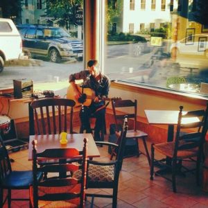 Acoustic guitarist plays in a sunlit cafe with wooden chairs and tables, large front windows reveal a street in Acadia National Park.