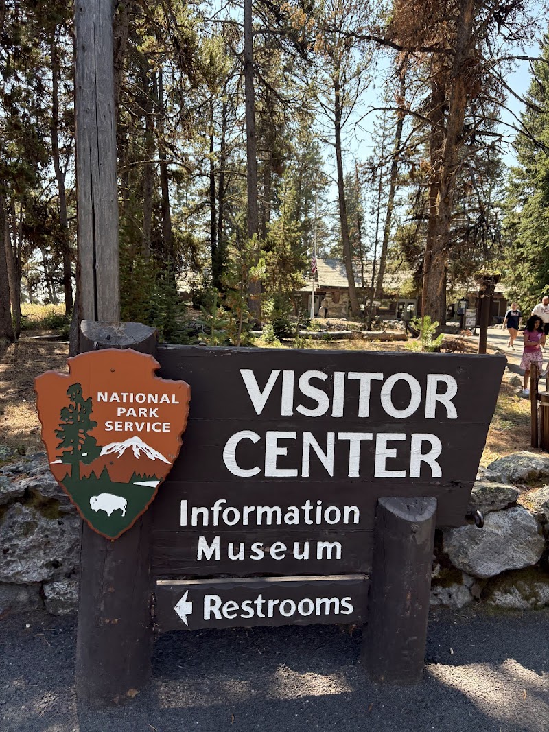 Yellowstone National Park sign and dark wooden Visitor Center board with Information, Museum, Restrooms, set among pines.