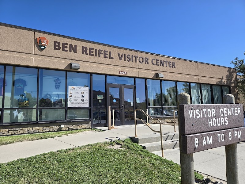 Brown brick visitor center with large glass windows and double doors, and a hours sign at Badlands National Park.