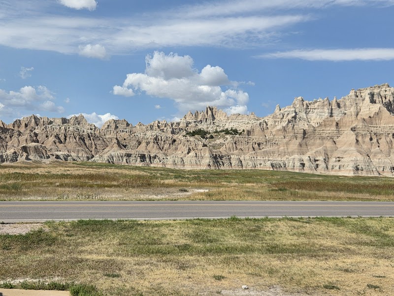 Wide view of eroded, layered badlands ridges across a grassy plain with a paved road in Badlands National Park.