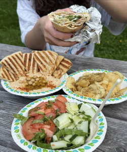 Acadia National Park picnic with a hand-held pita wrap, hummus plate, pita chips, and tomato-cucumber salad on a wooden table.
