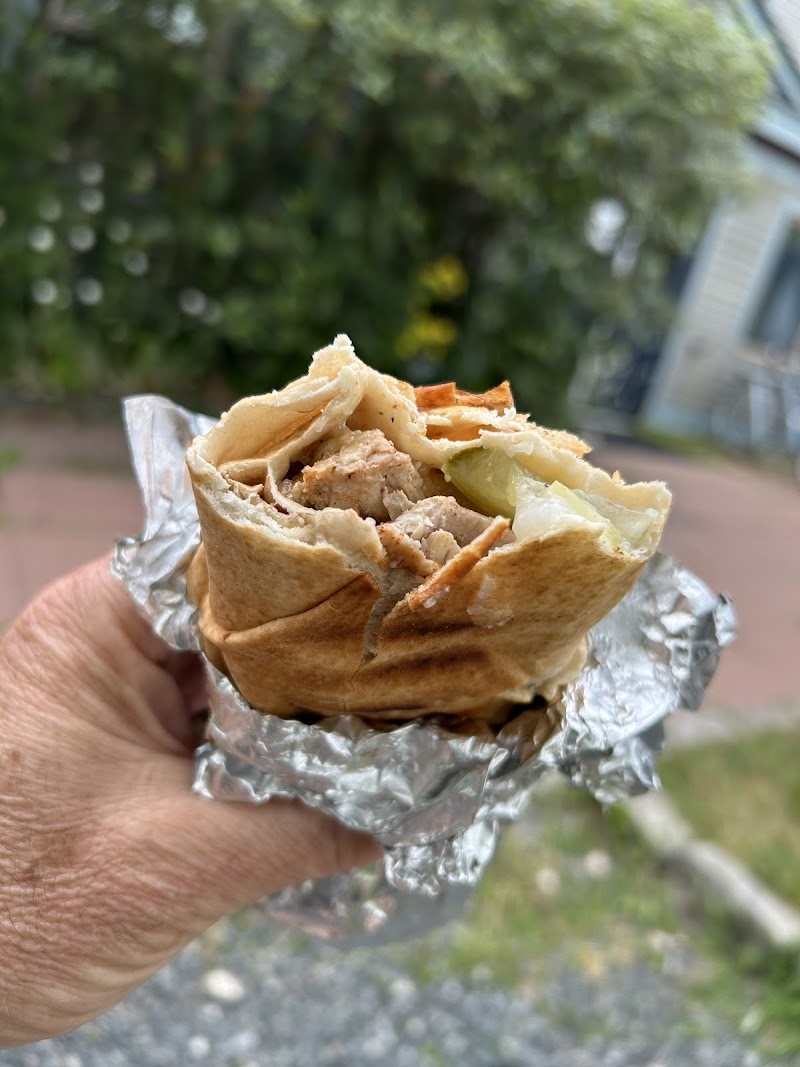 Hand holding a partially eaten pita wrap with grilled meat, onions, and pickles, wrapped in foil, outdoors in Acadia National Park.