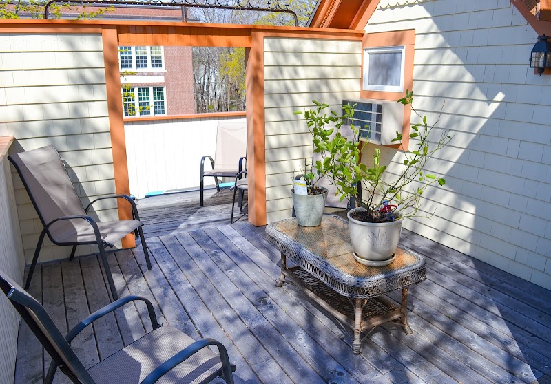 Cozy wooden deck with beige chairs and a wicker table holding potted plants, sunlit view in Acadia National Park.