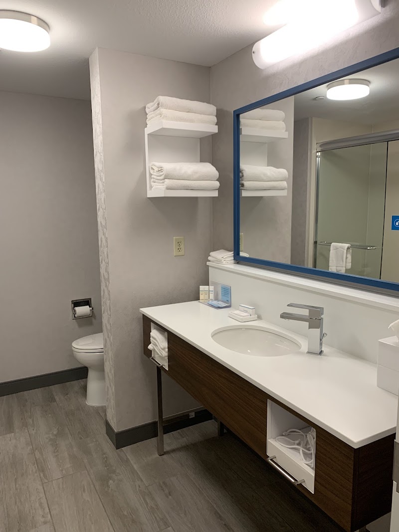 Modern hotel bathroom with white countertop sink, chrome faucet, blue-framed mirror, stacked towels on shelves, and a visible toilet in Arches National Park.