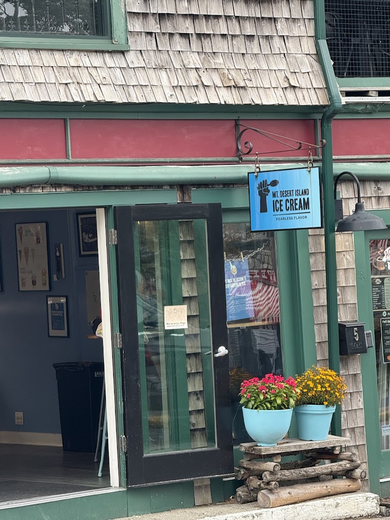 Exterior view of a rustic shop with green trim and shingle siding, blue pots of flowers and open glass doors in Acadia National Park.