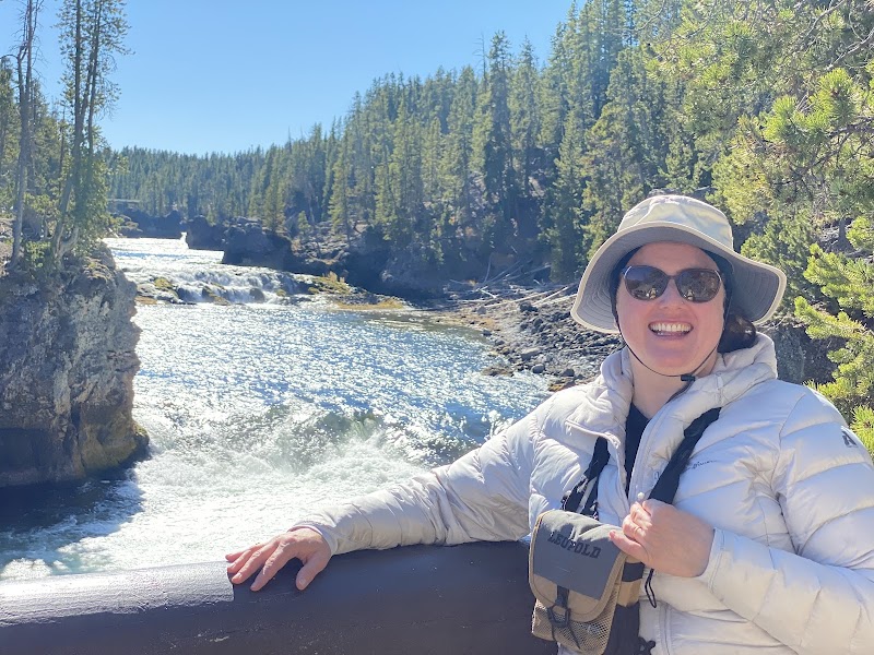 Smiling woman in a light jacket and hat leans on a railing beside the Yellowstone River at Lower Falls, Yellowstone National Park.