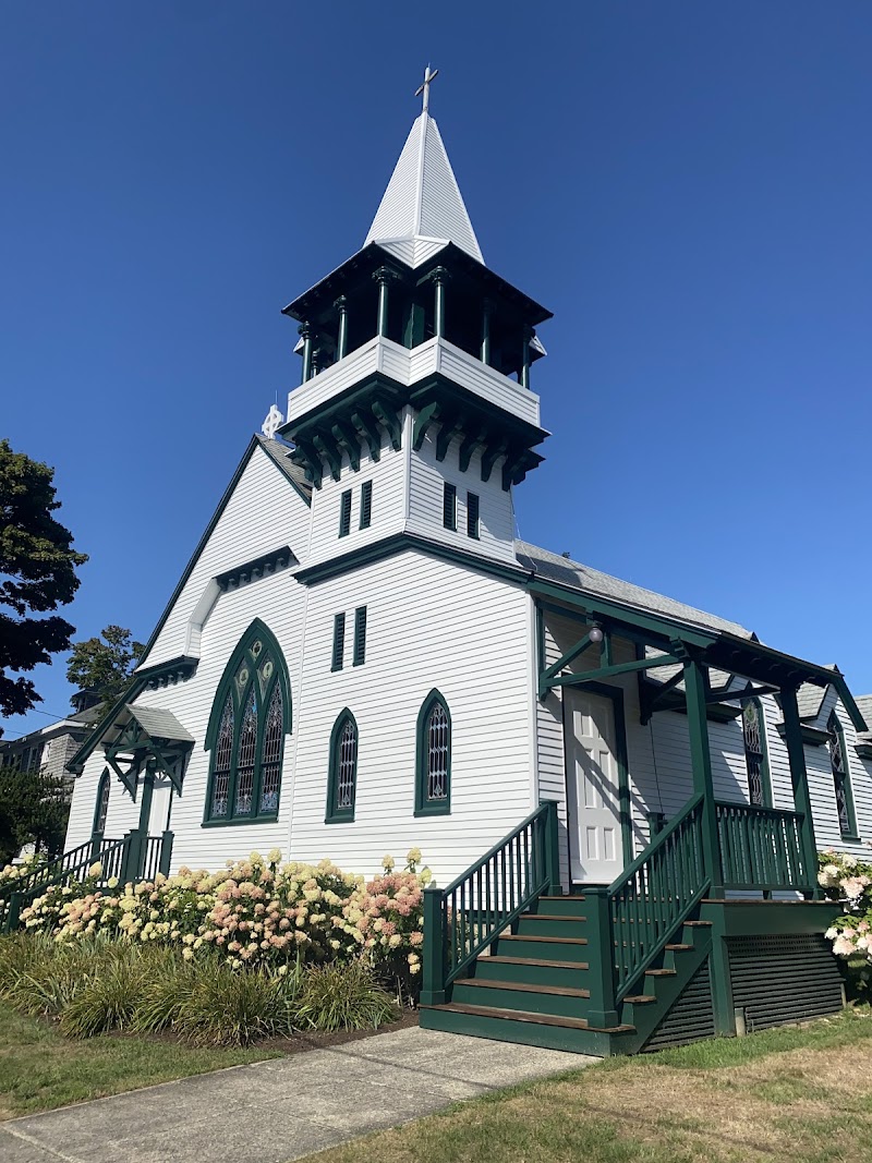 White wooden church with green trim and a tall steeple, green staircase, and hydrangeas under a blue sky in Acadia National Park.