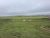 Wide grassy field in Badlands National Park with yellow wildflowers, three curved wooden shelters, a small building on the right, and distant low hills under a cloudy sky.