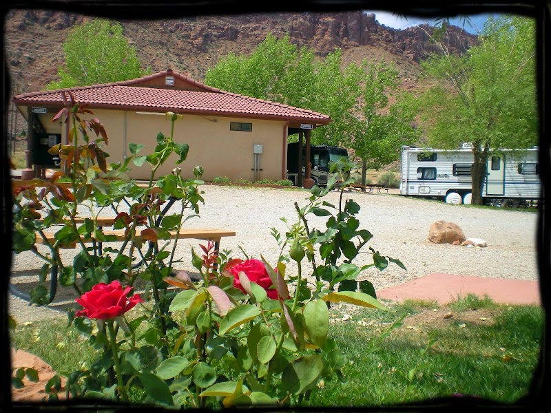 Roses and green trees frame a beige building with a red-tile roof and a parked RV at an Arches National Park campground.