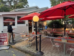 Outdoor seating area with red umbrellas, metal tables and chairs in front of a cafe, a man standing near the entrance, Acadia National Park.