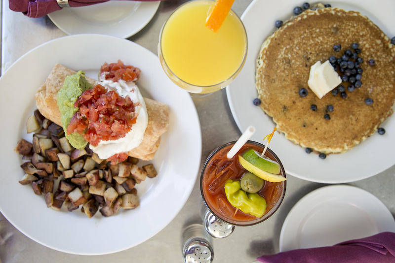 Acadia National Park cafe table with a plate of potato hash topped with crema, guacamole and pico, a blueberry pancake with butter, orange juice, and a lime-olive cocktail.