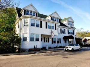 White three-story inn with dormers and black shutters on a street in Acadia National Park, with a small sign and a white car.