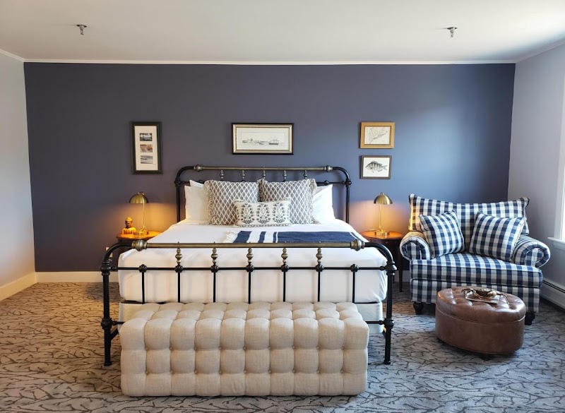 Acadia National Park lodge bedroom featuring a black metal bed, white linens, tufted bench, plaid chair, and navy wall art.