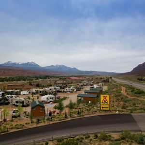 Desert campground filled with RVs and small cabins beside a paved road, near Arches National Park with distant snow‑capped mountains.