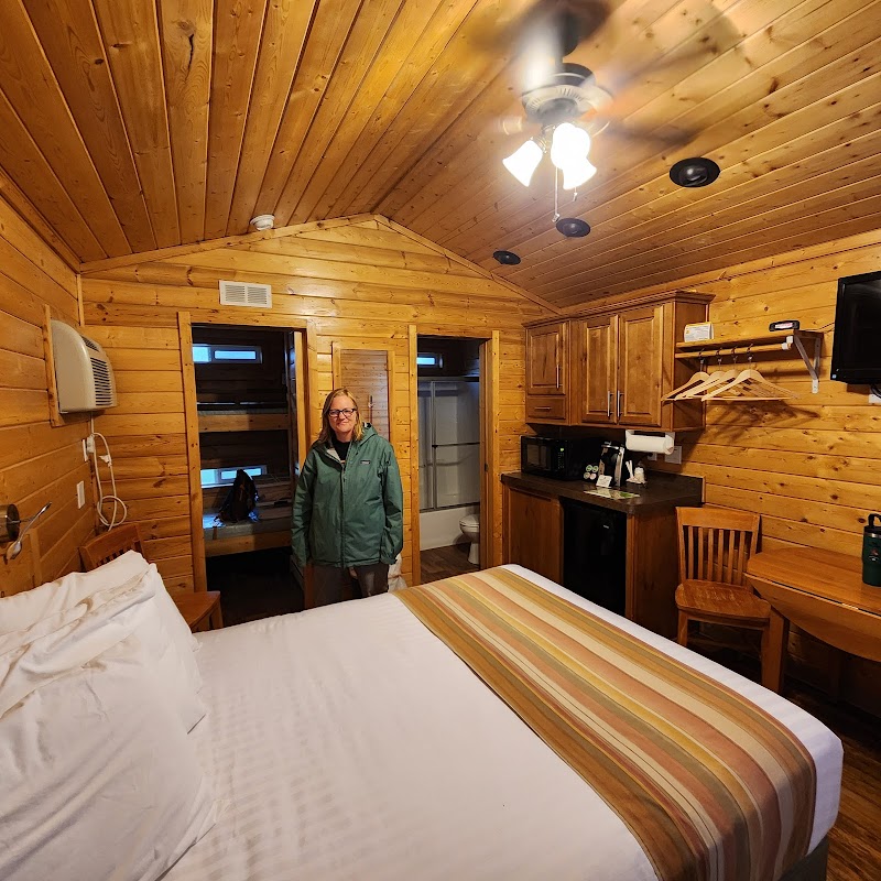 Wood-paneled cabin bedroom with a king bed, striped runner, kitchenette, and a woman in a green jacket near the doorway at Arches National Park.