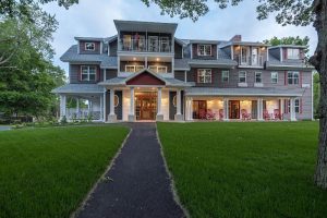 Three-story gray inn with red gabled entrance, wraparound porch, balconies, and lit windows in Acadia National Park.