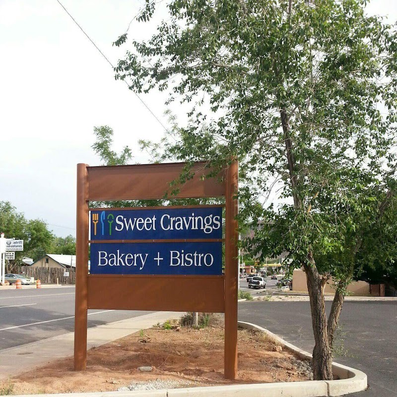 Brown wooden sign on a dirt patch beside a road, blue panels advertise a bakery and bistro near Arches National Park.