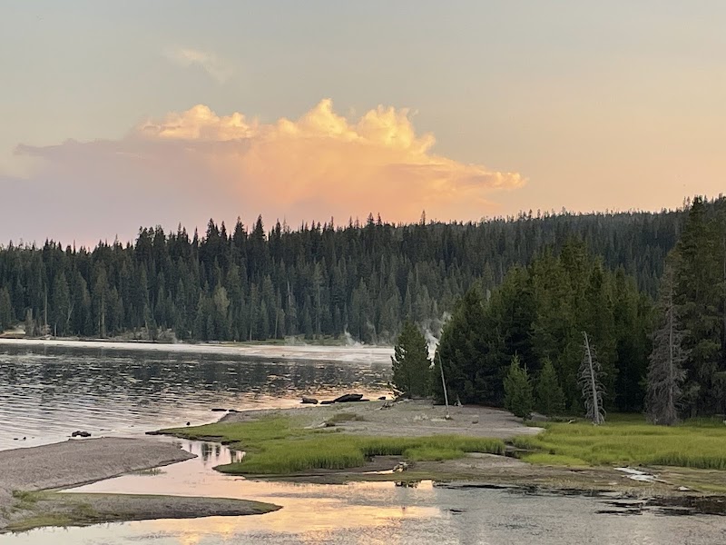 Sunset over Yellowstone Lake in Yellowstone National Park, calm water reflecting orange clouds beside a pine shoreline.