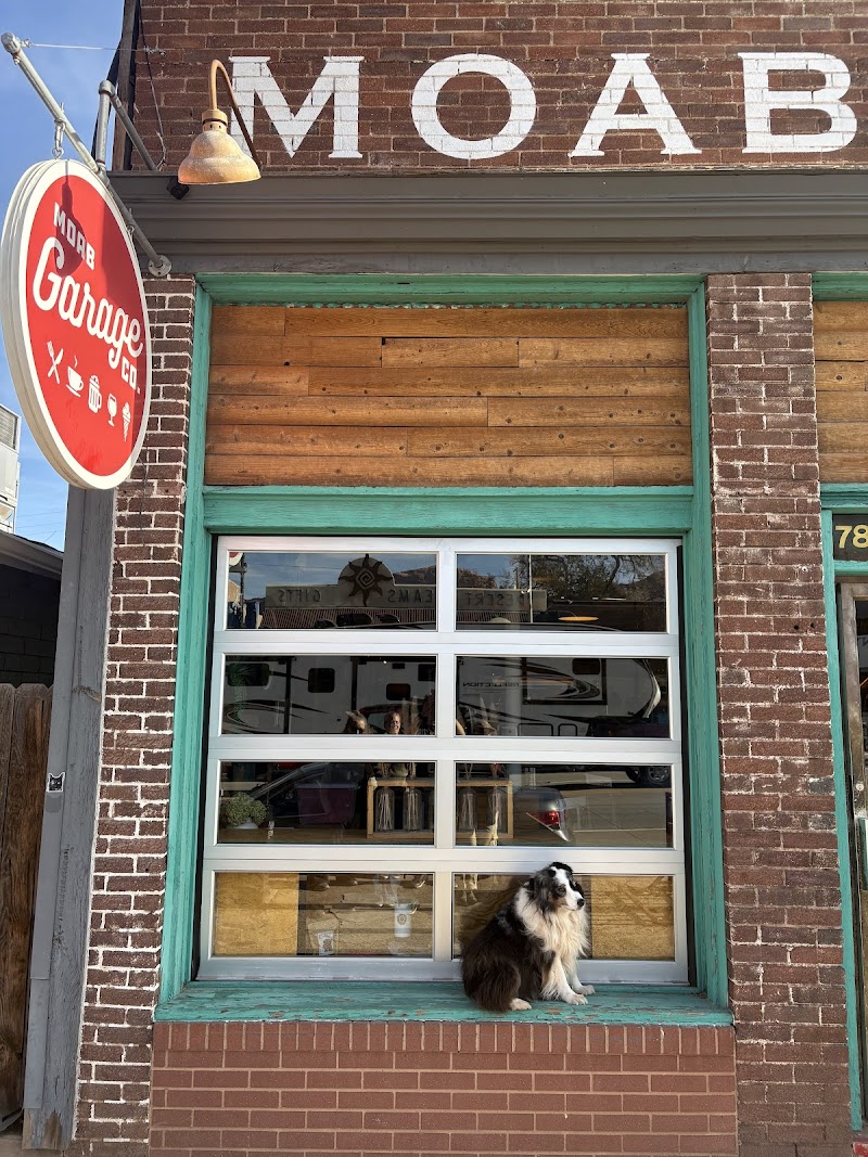 Brick storefront with green trim and a large glass door; a dog sits on the window ledge in Arches National Park.