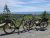 Two electric bicycles with pannier bags parked on a gravel overlook in Acadia National Park, with forested hills and the blue Atlantic beyond.