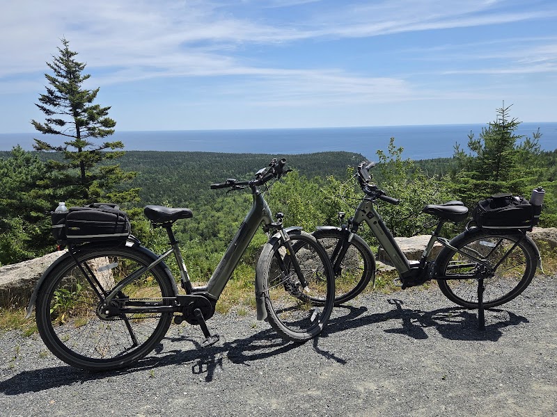 Two electric bicycles with pannier bags parked on a gravel overlook in Acadia National Park, with forested hills and the blue Atlantic beyond.