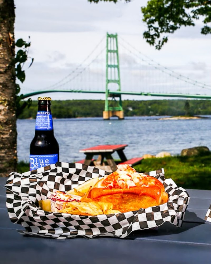 Lobster roll and beer on a picnic table with a distant green bridge and waterfront scenery in Acadia National Park.