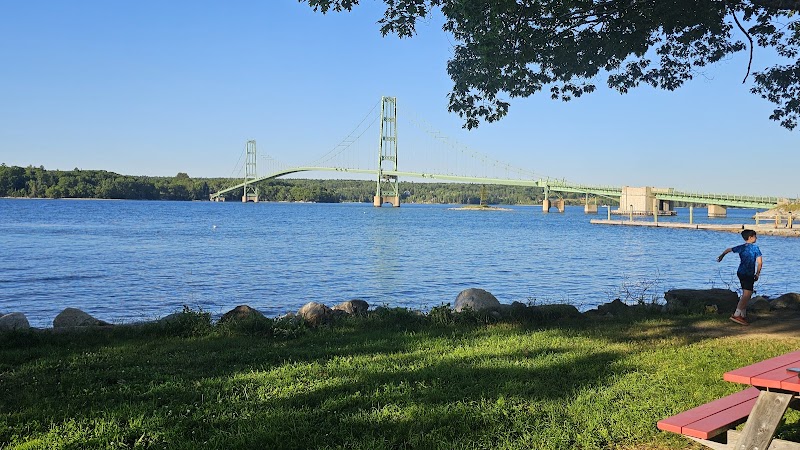 Bridge over blue water at Acadia National Park, with grassy shore, rocky edge, and a person near a red picnic table.