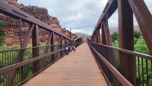 Arches National Park: long brown metal truss bridge with railings, three hikers, red cliffs and green desert foliage under a cloudy sky.