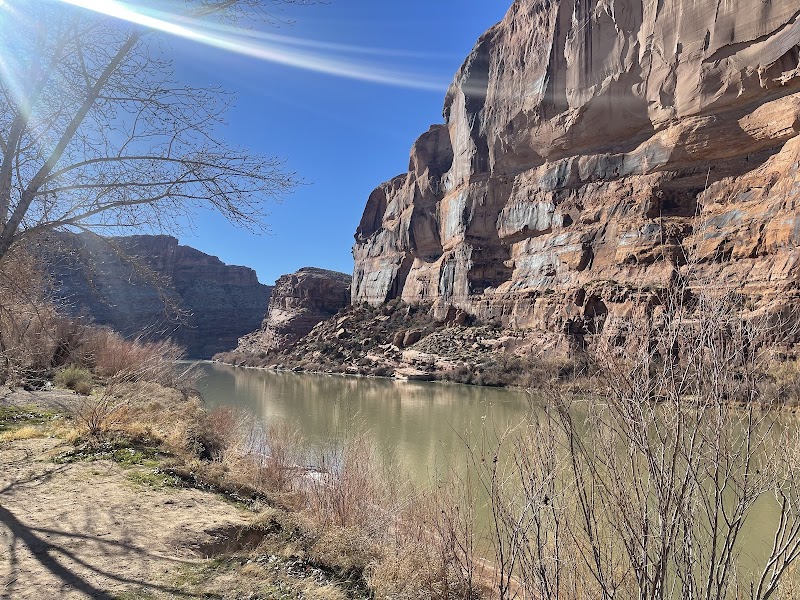 Arches National Park scene with a calm river beside towering red rock cliffs and leafless shrubs along a dirt trail.