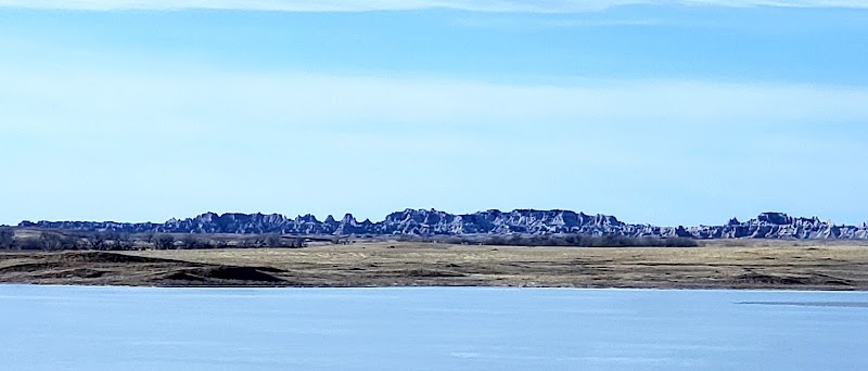 Calm lake in the foreground with a flat grassland plain and jagged Badlands ridges across the horizon in Badlands National Park.