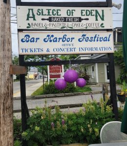 Two stacked signs advertise a bakery and Bar Harbor Festival, with purple lanterns and green plants along a street in Acadia National Park.