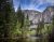 Yosemite Valley scene with a tall granite cliff and a slender waterfall to the right, framed by pine trees and a calm Merced River.