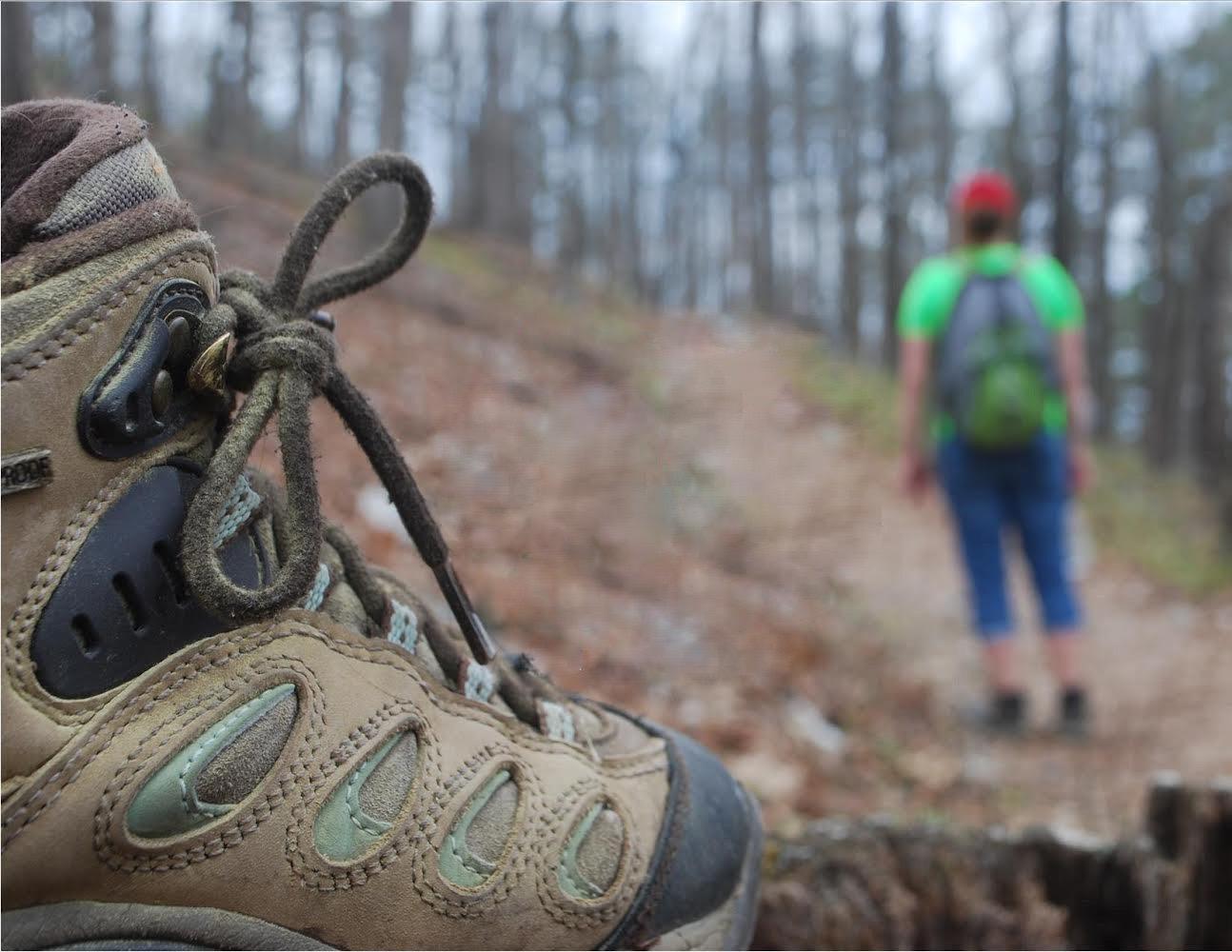 Close-up of a rugged hiking boot on a leaf-covered forest trail, with a hiker in a bright green shirt and backpack ahead in Hot Springs National Park.