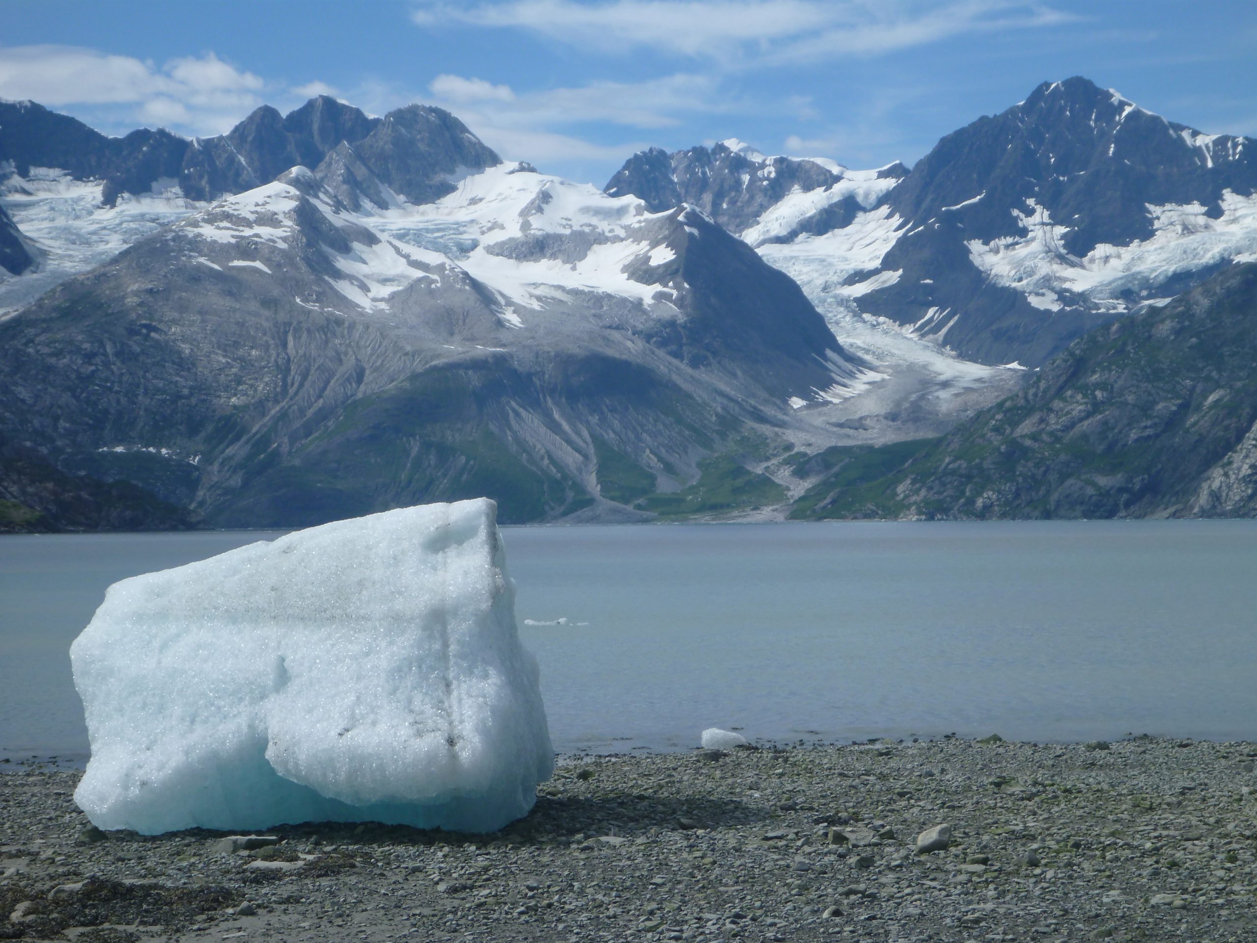 Iceberg rests on a pebbled shore with snow‑capped, rugged peaks and glaciers across Glacier Bay National Park & Preserve, Alaska.