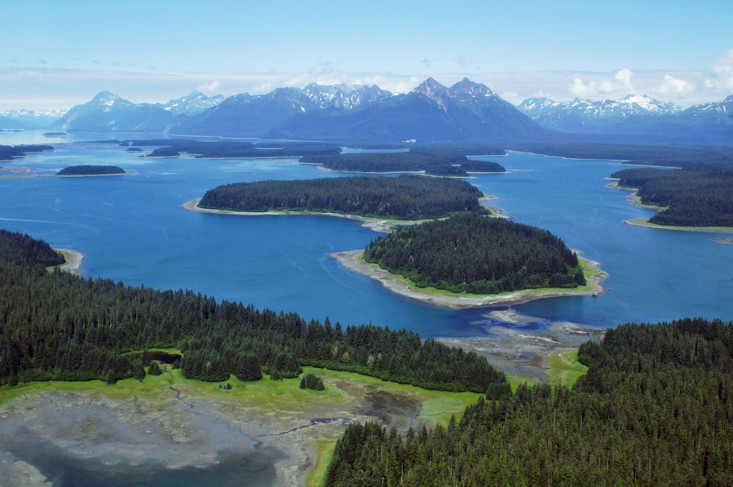 Aerial view over Glacier Bay National Park & Preserve with forested islands, inlets, and snow‑capped mountains under a blue sky.