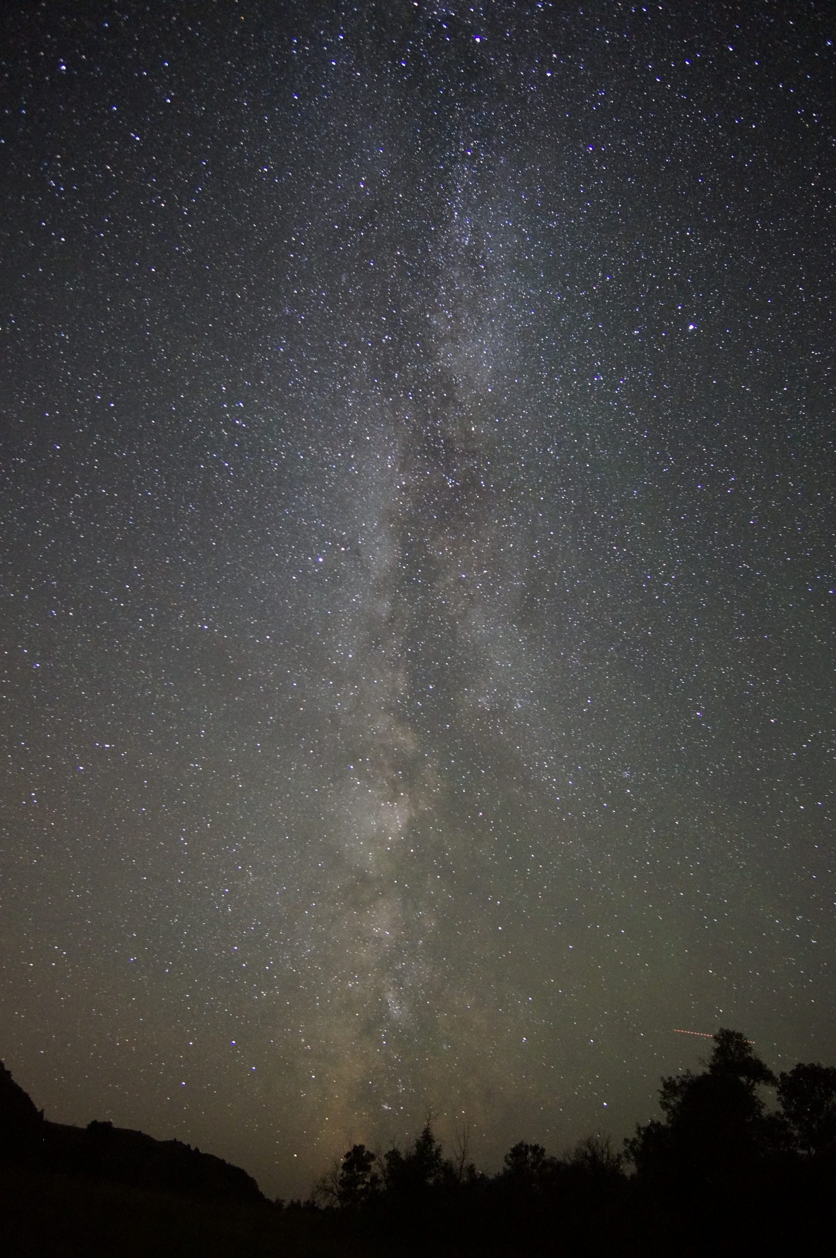 Milky Way stretches across the night sky over Theodore Roosevelt National Park's badlands, with dark silhouettes of trees on the horizon.
