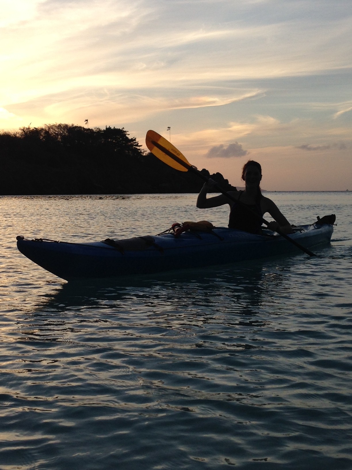 A silhouette of a person paddling a blue kayak at sunset near a rocky, tree-covered island in Virgin Islands National Park.