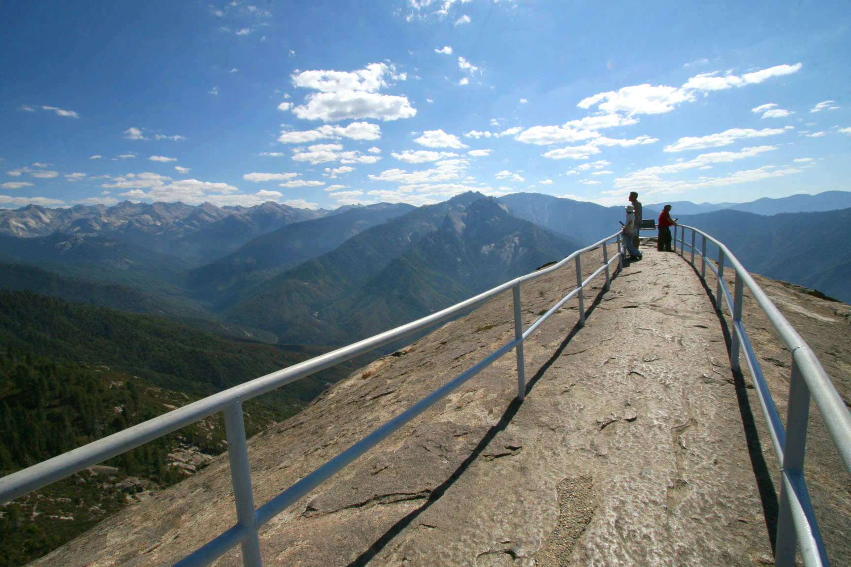 Visitors stand on a paved, railing-lined viewing platform on Moro Rock, Sequoia & Kings Canyon National Parks, with Sierra Nevada peaks and a blue sky.