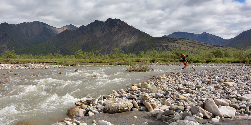 Two hikers cross a rocky riverbank with glacial mountains rising behind in Gates of the Arctic National Park & Preserve.