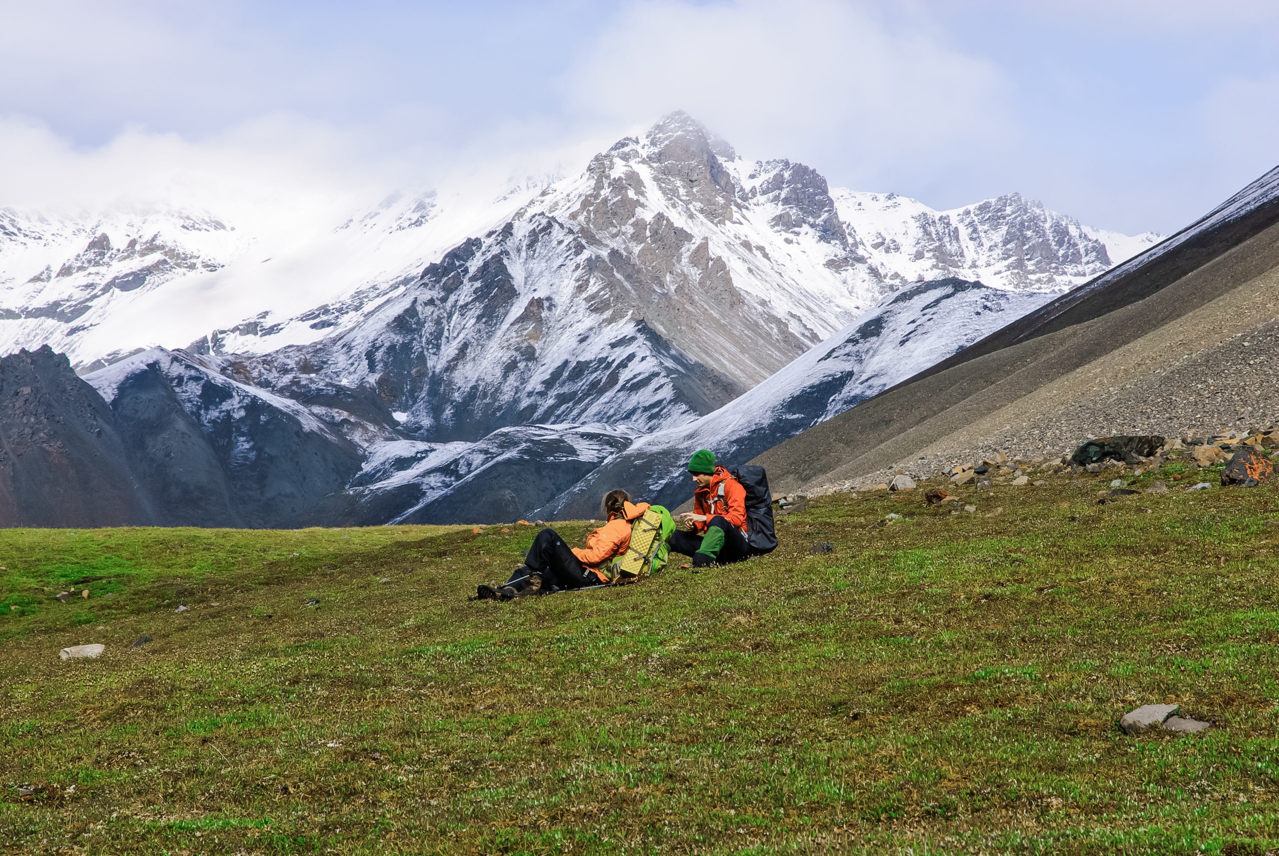 Two hikers resting on a grassy alpine meadow with snow‑capped Wrangell mountains in Wrangell–St. Elias National Park.