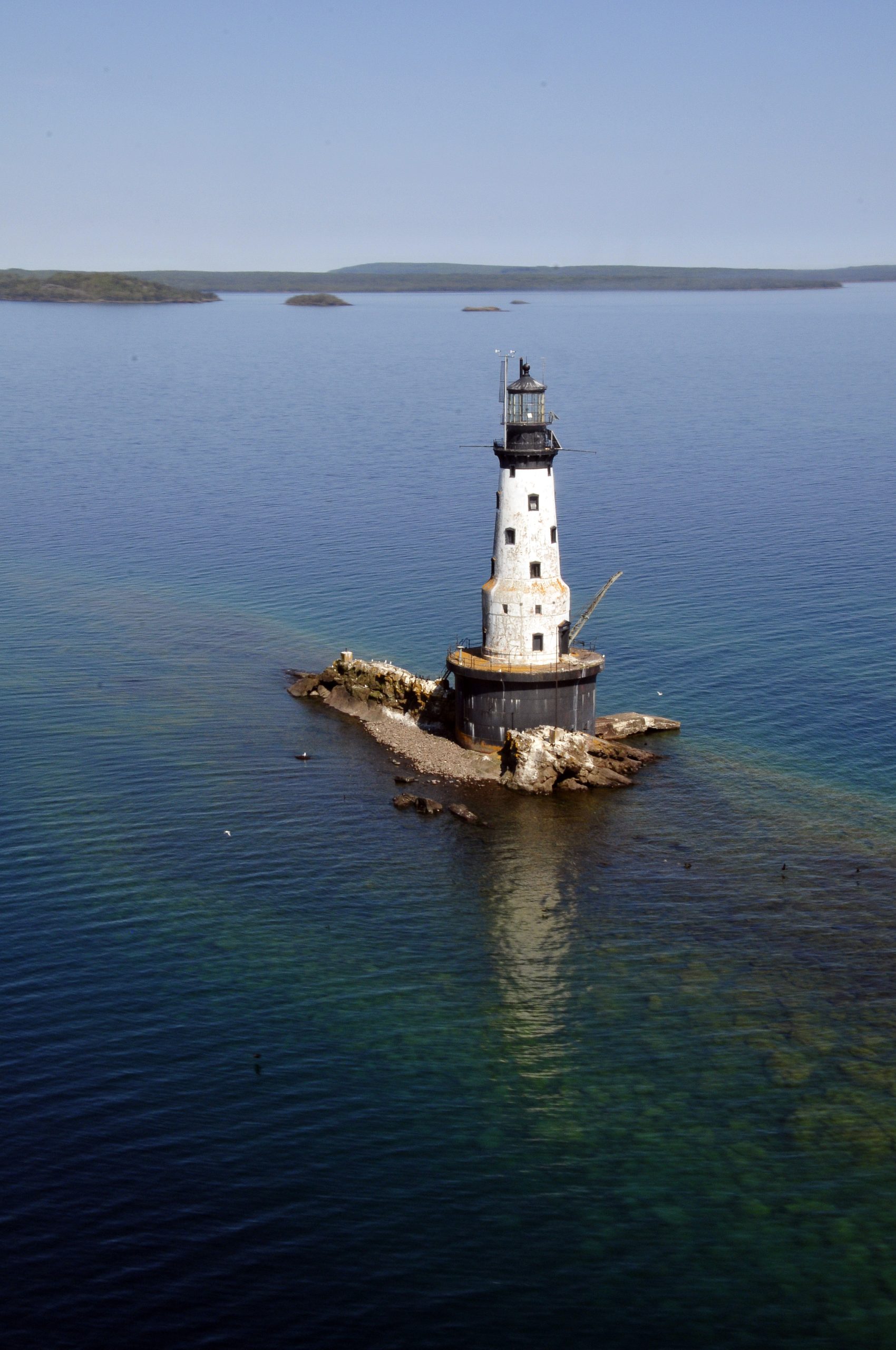 Isle Royale Lighthouse stands on a rocky islet in calm blue waters, with a tall white tower and black lantern.