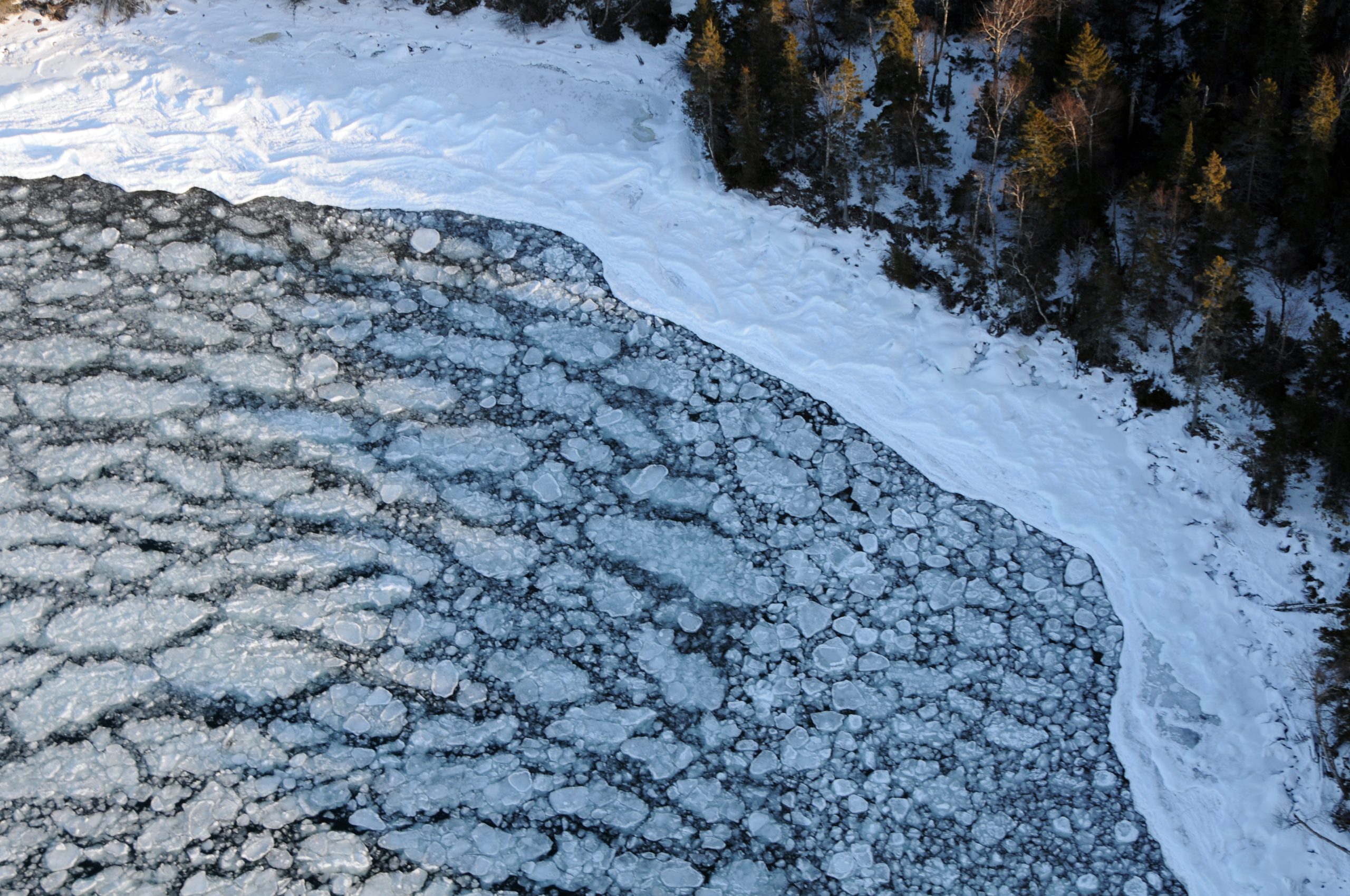 Aerial view of jagged ice floes along a snow-covered shoreline with a forested ridge of trees nearby, Isle Royale NP.