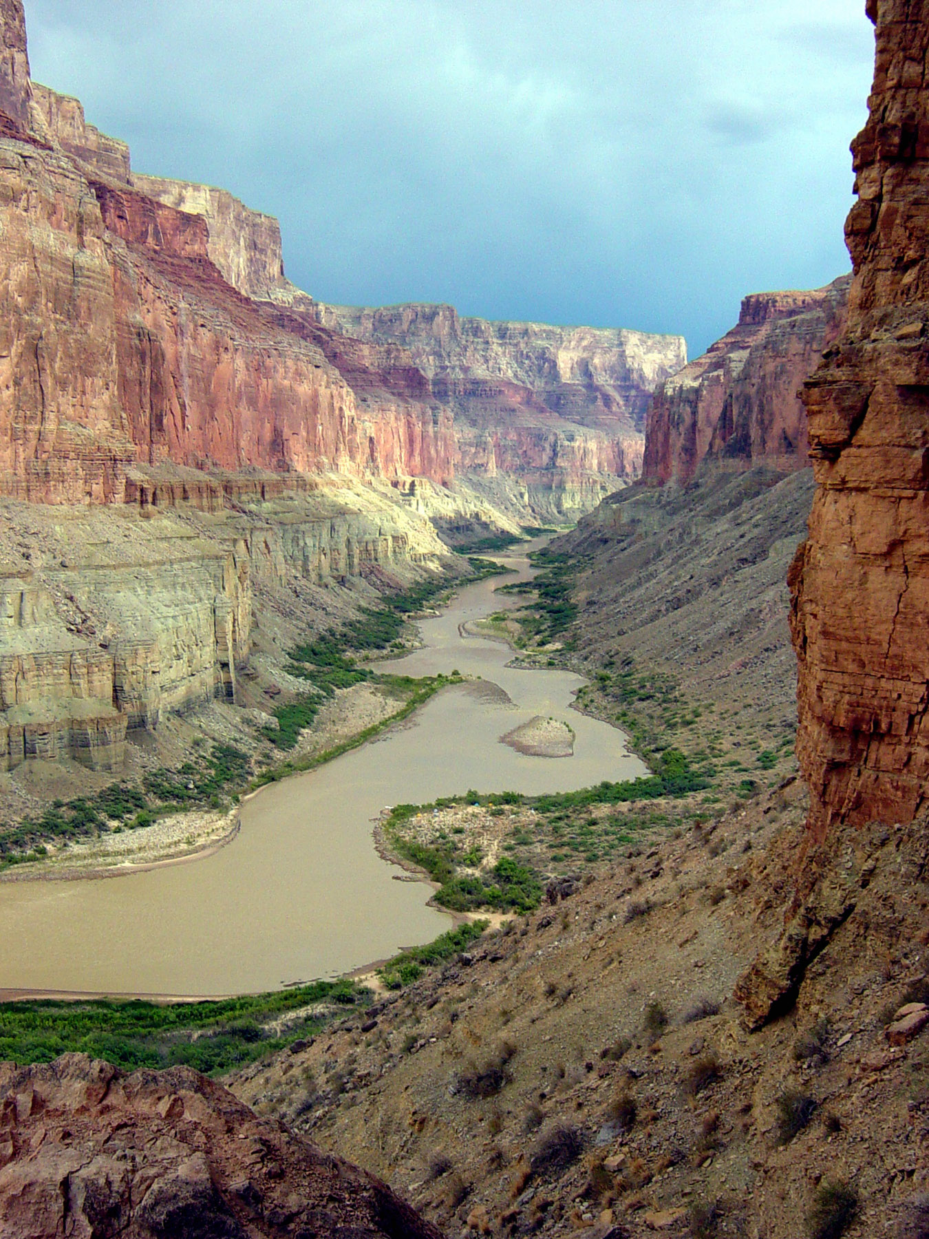 Colorado River winds through the Grand Canyon National Park, slicing between towering red rock walls under a moody sky.
