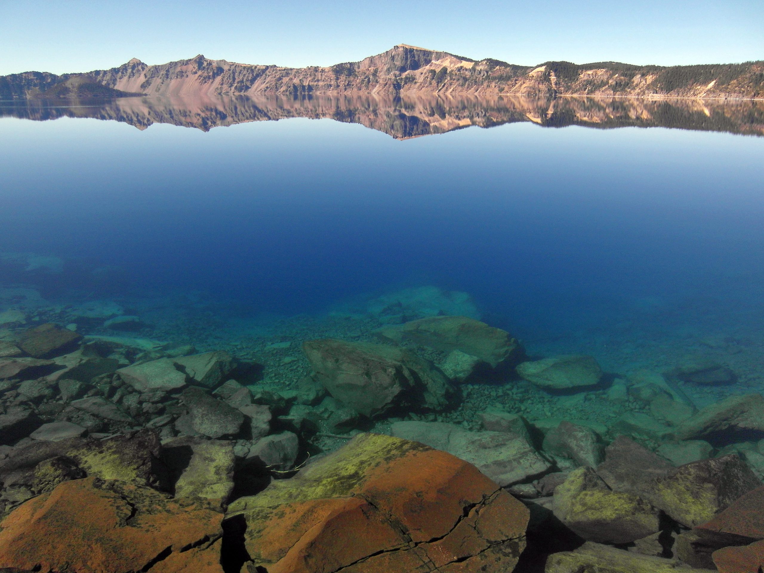 Crater Lake shoreline at Crater Lake National Park with clear blue water and a rocky foreground along the calm lake surface.