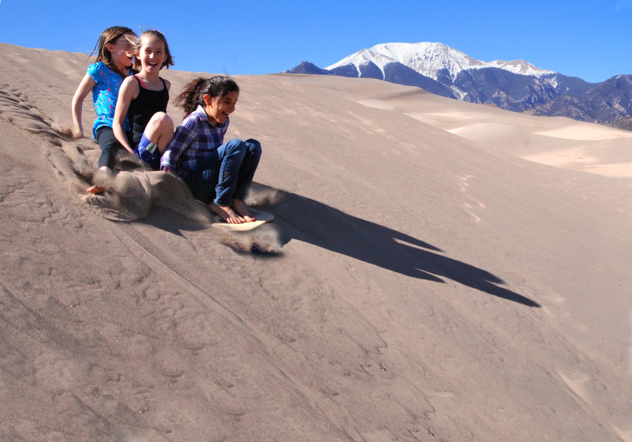 Three kids ride a sandy dune at Great Sand Dunes National Park & Preserve, with snow-capped mountains rising in the distance against a clear blue sky.