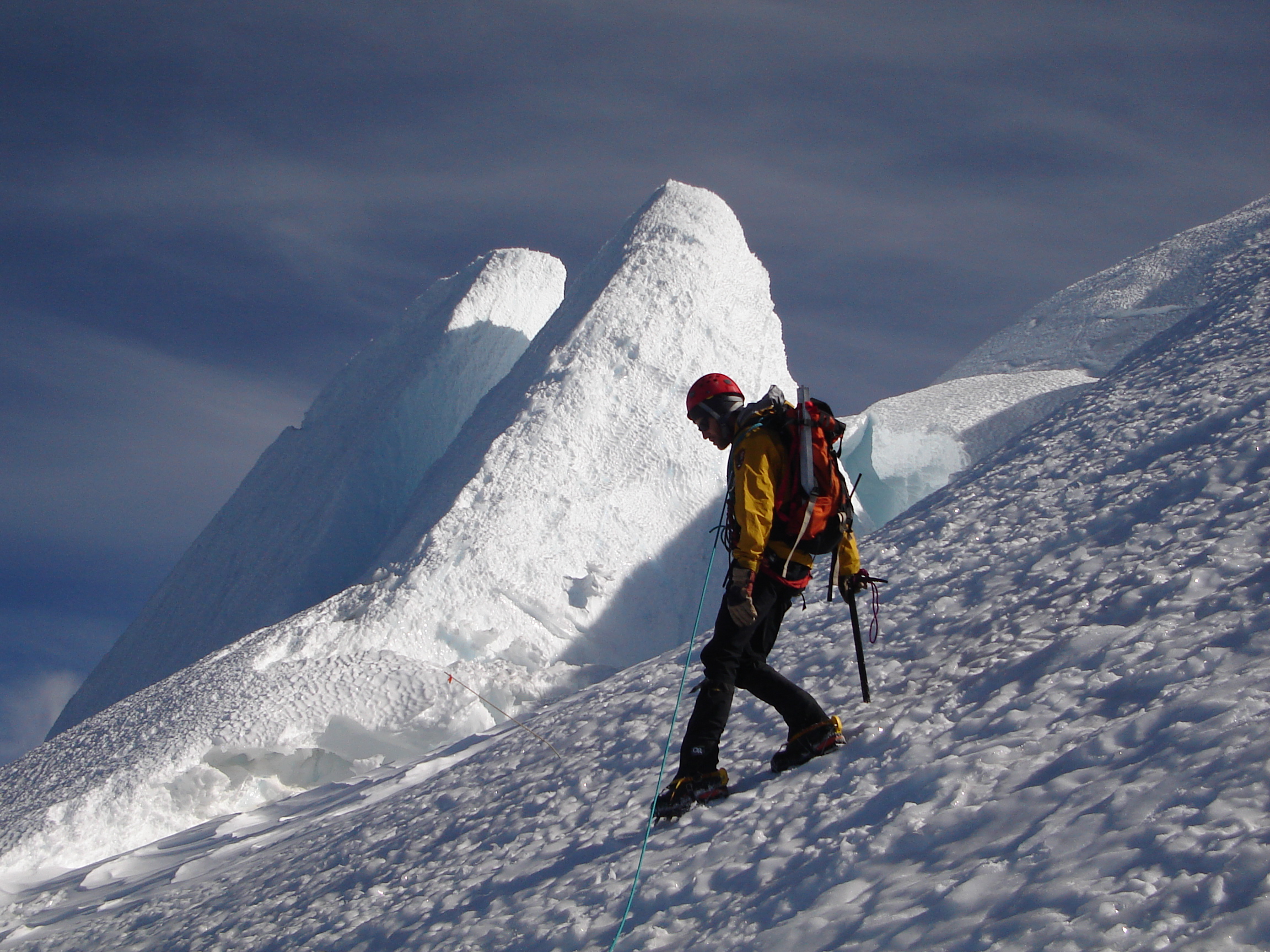 Climber in a yellow jacket and red helmet ascends a snowy slope with a jagged icy peak in Mount Rainier National Park.